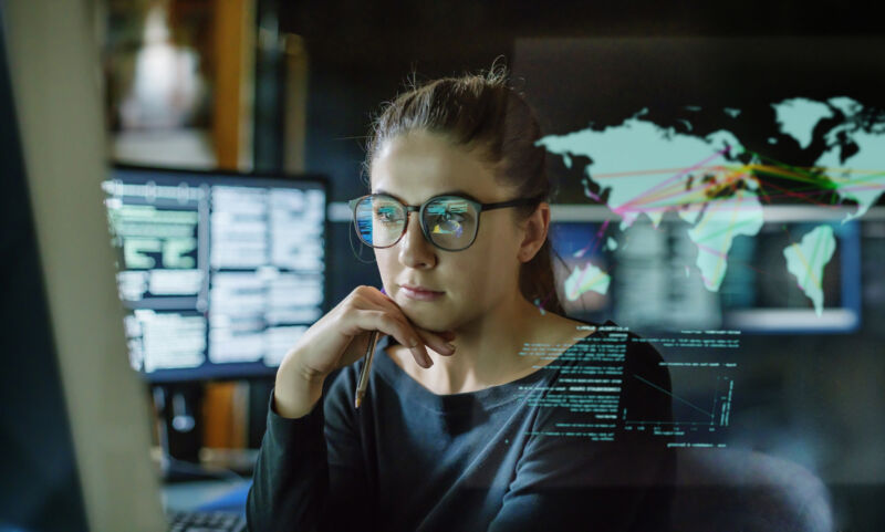 Imagen de archivo de una mujer joven con anteojos rodeada de monitores de computadora en una oficina oscura.  Frente a ella hay una pantalla transparente que muestra un mapa del mundo con algunos datos.